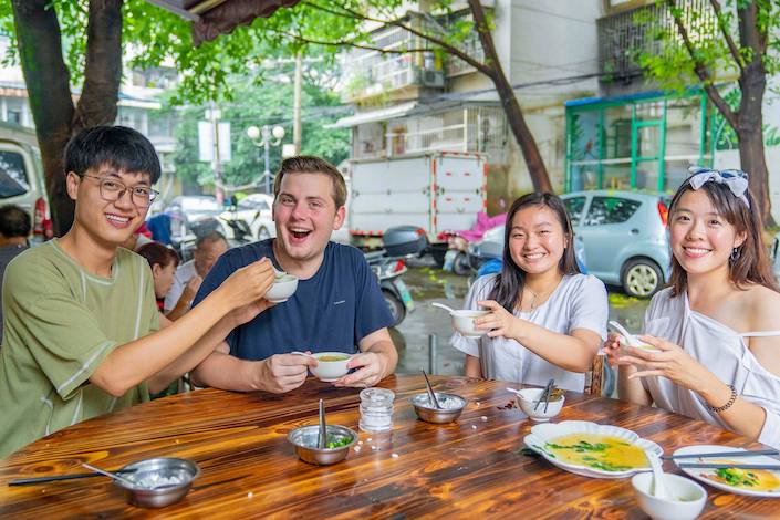 a CLI student eats a meal outdoors beside a road with three Chinese people