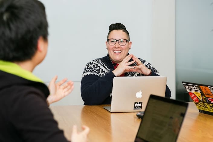 a woman with short hair sits at a table with an Apple laptop in front of her and smiles while listing to a man sitting across from her