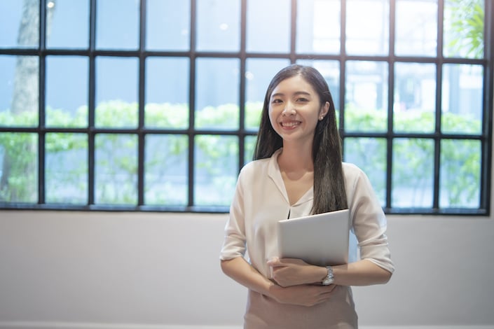 a smiling Chinese woman with long hair standing in front of a window holding some papers