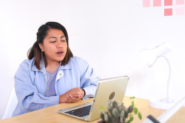 a woman in a blue shirt with clasped hands speaks while looking at a laptop on the table in front of her