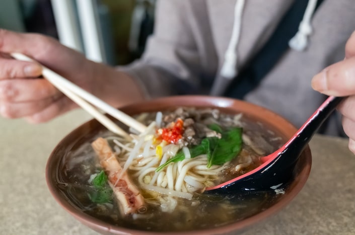 two hands holding a pair of chopsticks and a spoon while eating a bowl of Chinese lamb soup with noodles